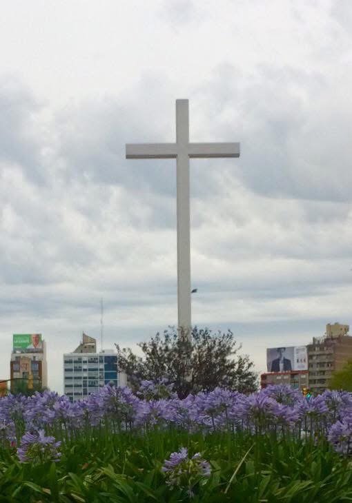La cruz blanca de Tres Cruces elevándose sobre los edificios, con agapantos violetas en primer plano y un cielo nublado.